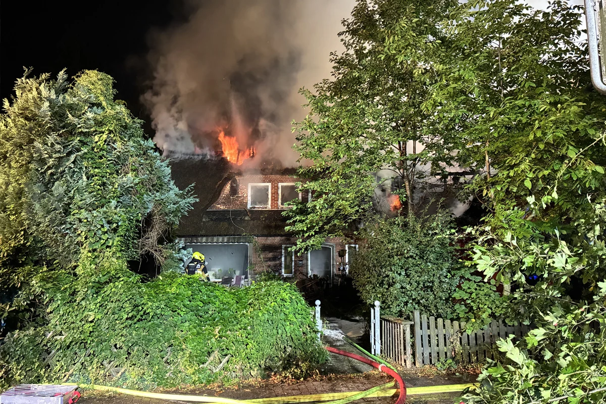 Ein Wohnhaus steht in Flammen, deutlich sichtbar sind Flammen und Rauch auf dem Dach. Ein Feuerwehrmann ist im Einsatz vor dem Gebäude zu sehen.