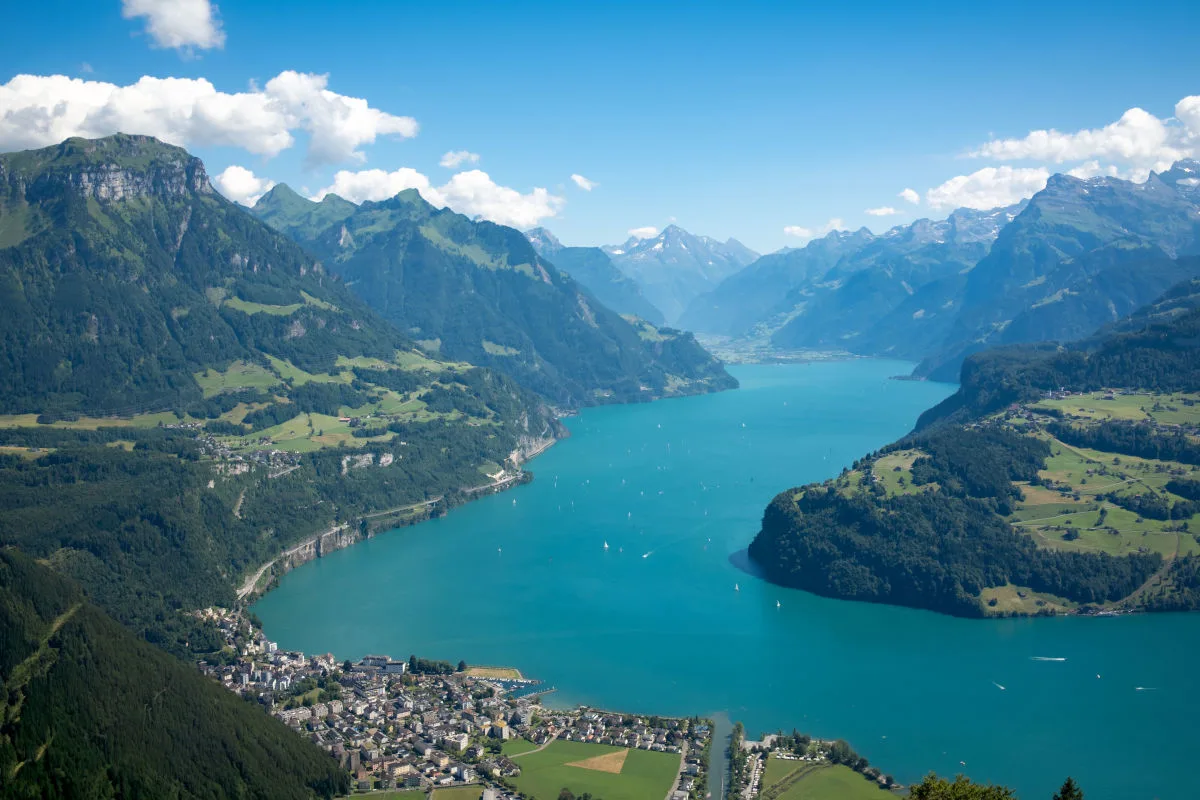 Die Luftaufnahme zeigt den türkisglänzenden Vierwaldstättersee, umrahmt von grünen Bergen, unter einem klaren, blauen Himmel. Im Vordergrund ist eine kleine Ortschaft erkennbar, flankiert von Wiesen und Bäumen.
