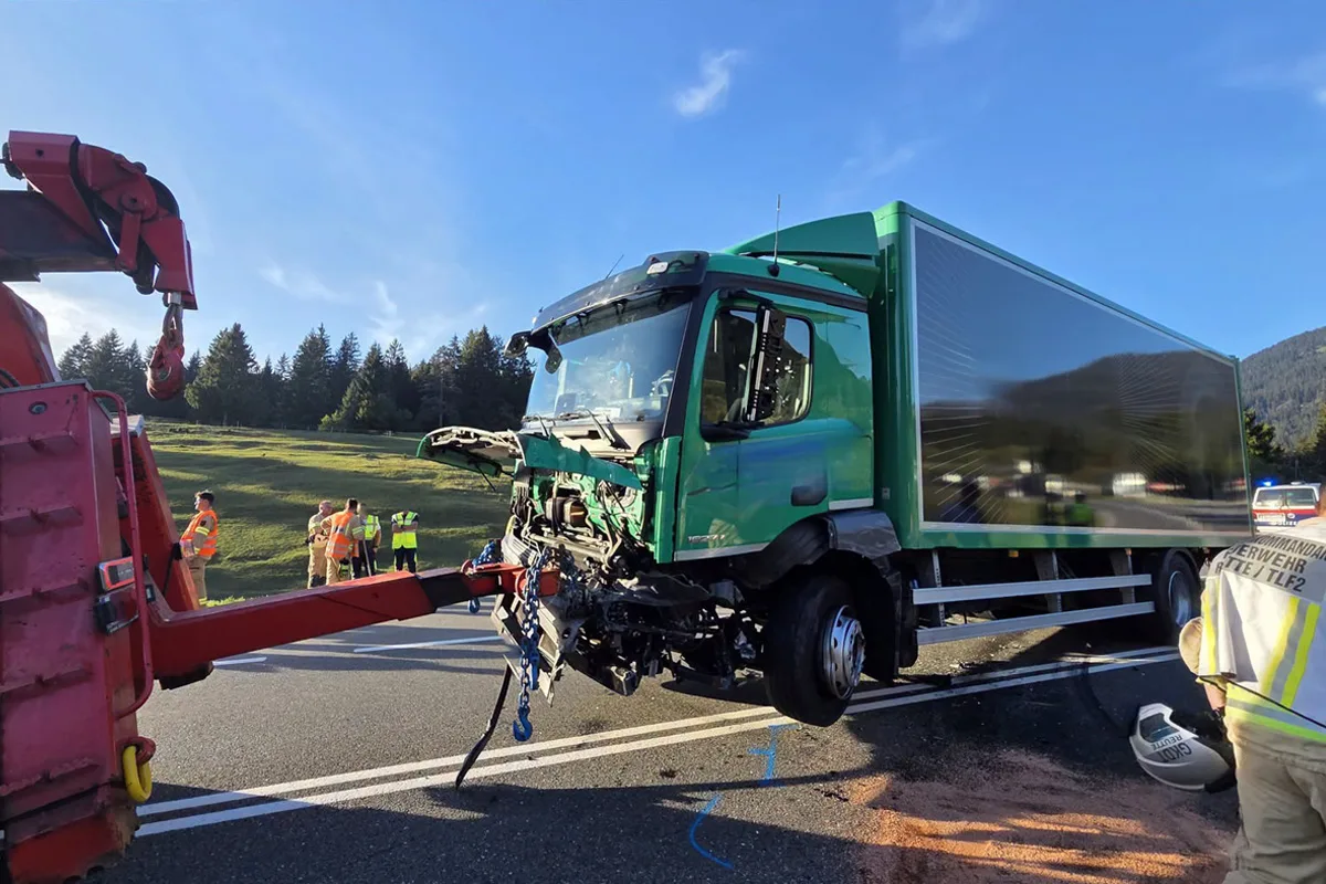 Ein grüner Lastwagen mit beschädigter Front wird von einem roten Abschleppfahrzeug auf einer Straße abgeschleppt. Im Hintergrund stehen Personen in Warnwesten.