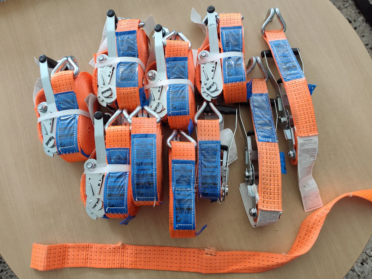 Auf einem Tisch liegen mehrere orangefarbene Zurrgurte mit blauen Etiketten. Die Gurte sind eng zusammengerollt und mit Metallratschen ausgestattet.