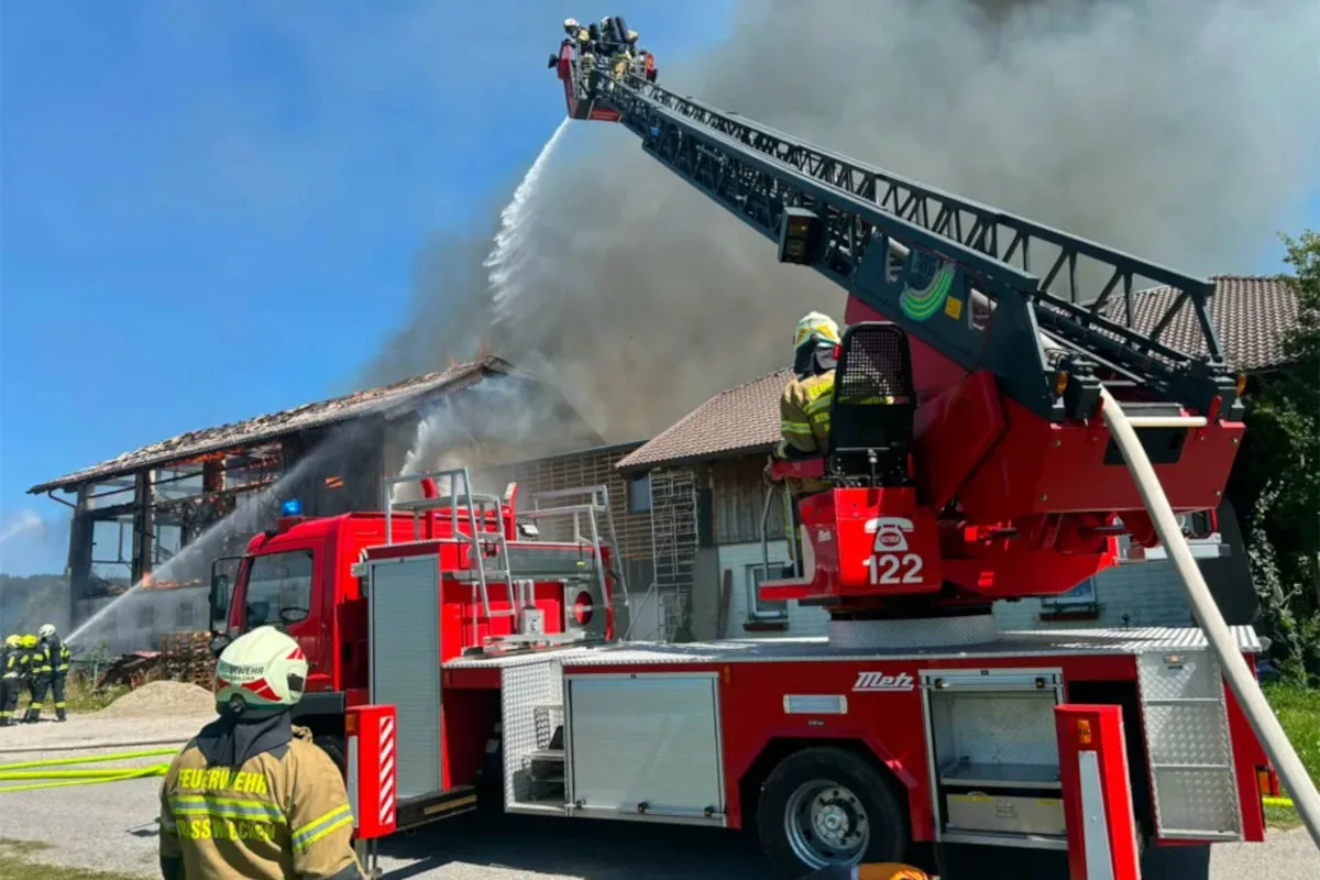 Ein Feuerwehrmann nutzt eine Drehleiter, um das Dach eines brennenden Gebäudes zu löschen. Im Hintergrund sind Rauch und beschädigte Gebäudeteile sichtbar.