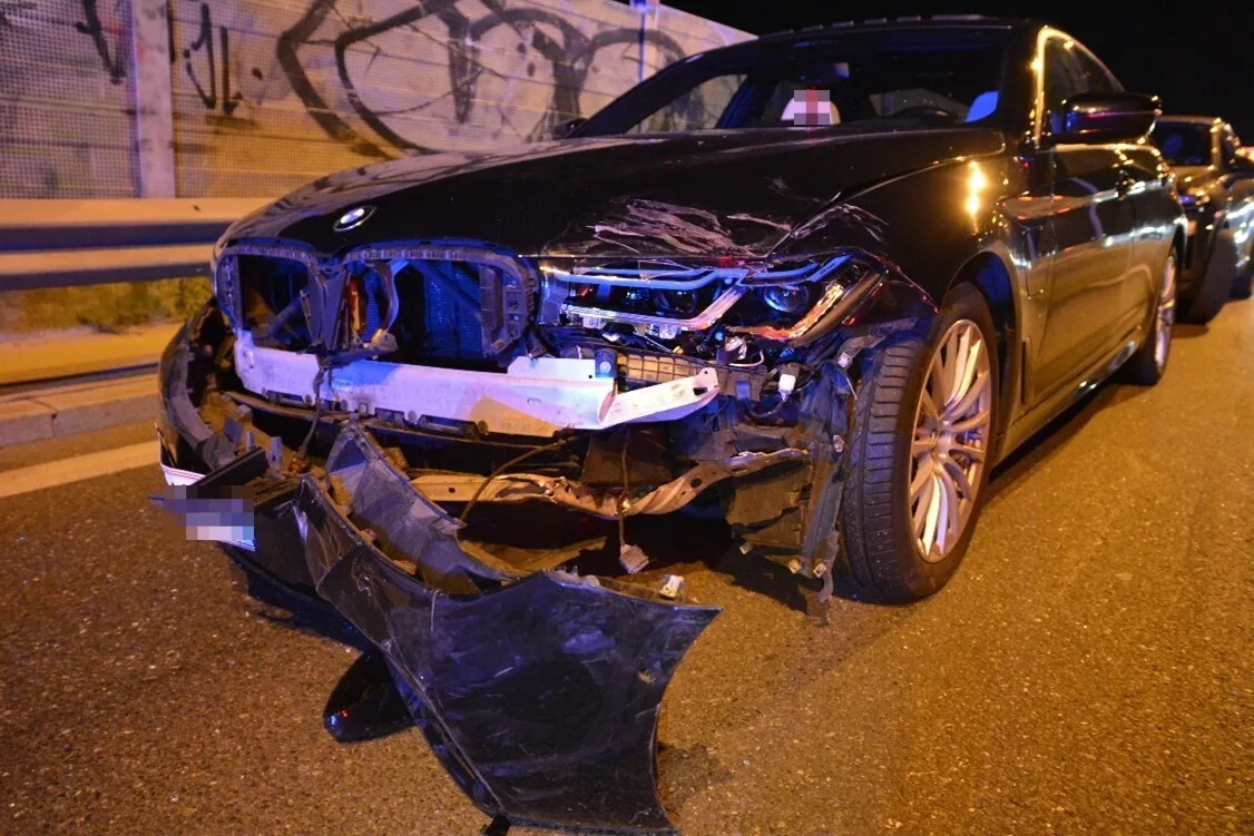Ein schwarzer BMW steht stark beschädigt auf einer Straße; der Frontbereich des Fahrzeugs ist stark eingedrückt und abgerissen.
