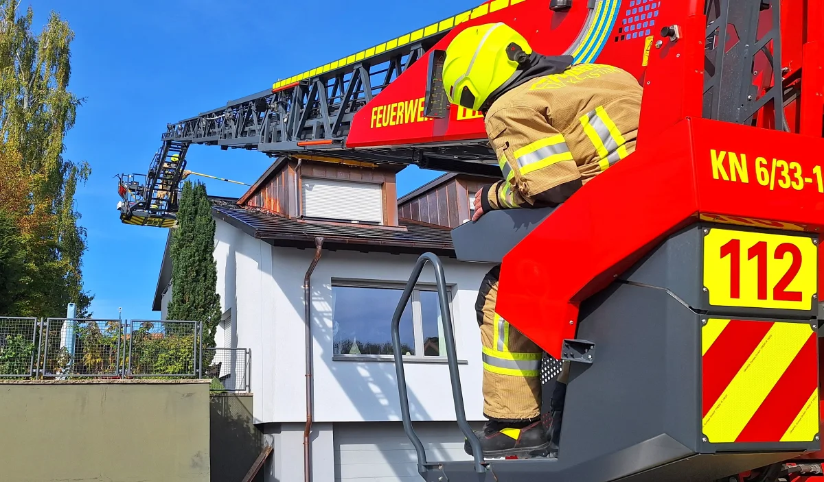 Ein Feuerwehrmann in Schutzausrüstung steht auf einer Drehleiter, um einen Dachbrand bei einem Wohnhaus zu bekämpfen. Flammen sind auf dem Dach sichtbar.