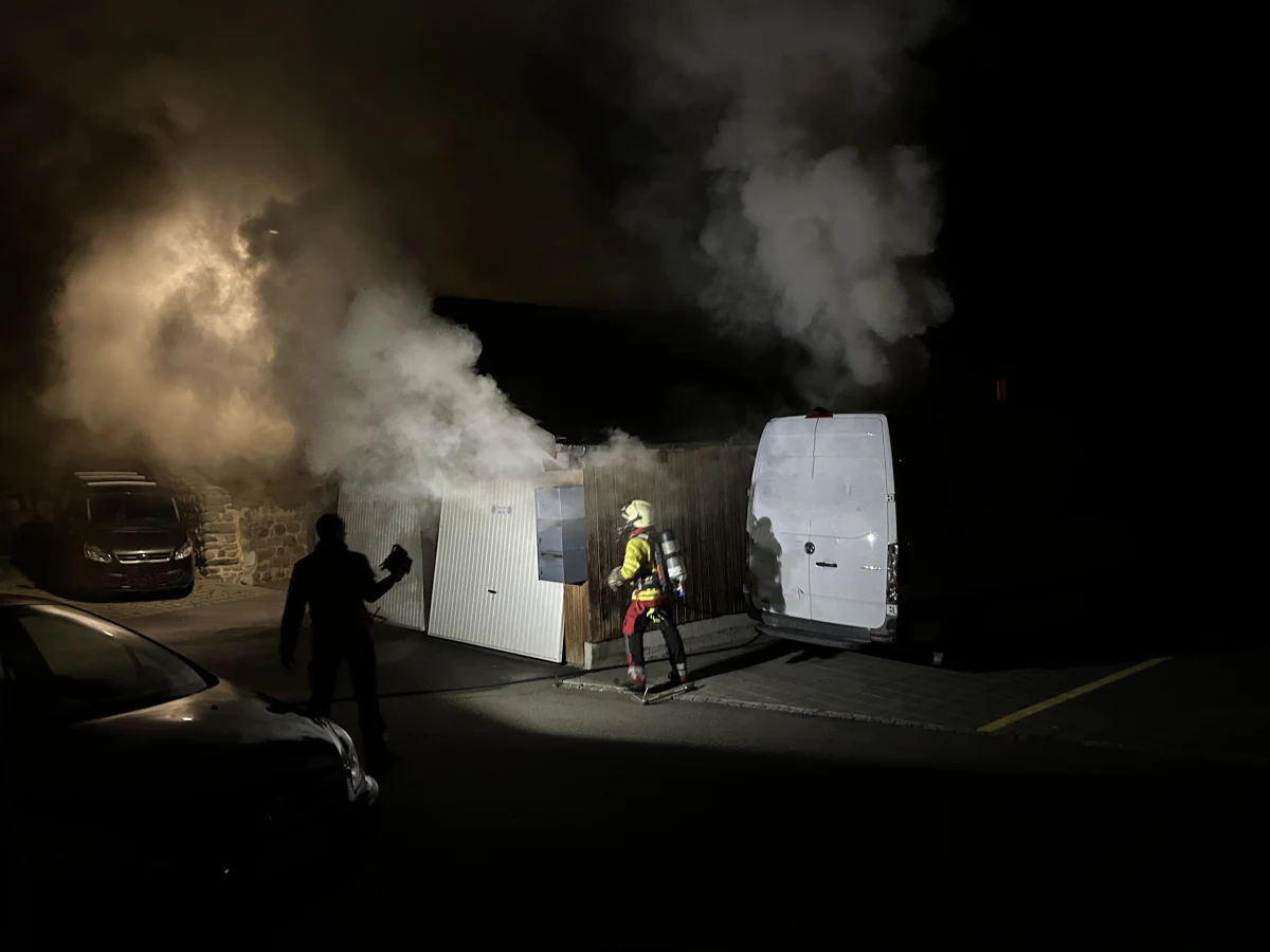 Ein Feuerwehrmann in voller Einsatzkleidung steht vor einer rauchenden Garage im Dunkeln. Ein weißer Lieferwagen ist daneben geparkt, und Rauch steigt auf.