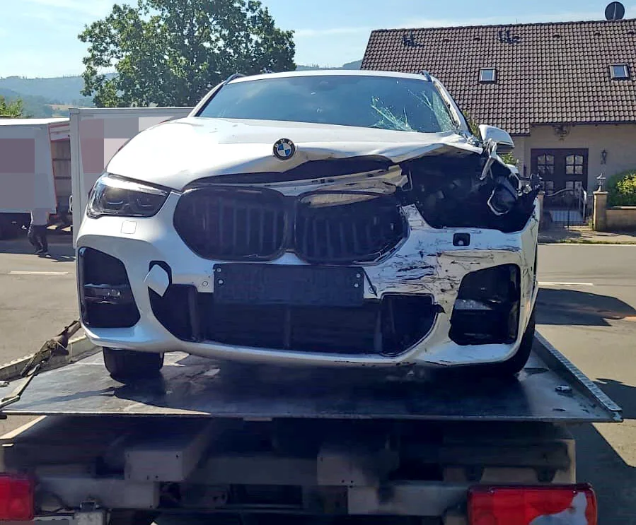 Ein weißer BMW mit erheblichem Frontschaden steht auf einem Abschleppwagen. Im Hintergrund sind Gebäude sichtbar.