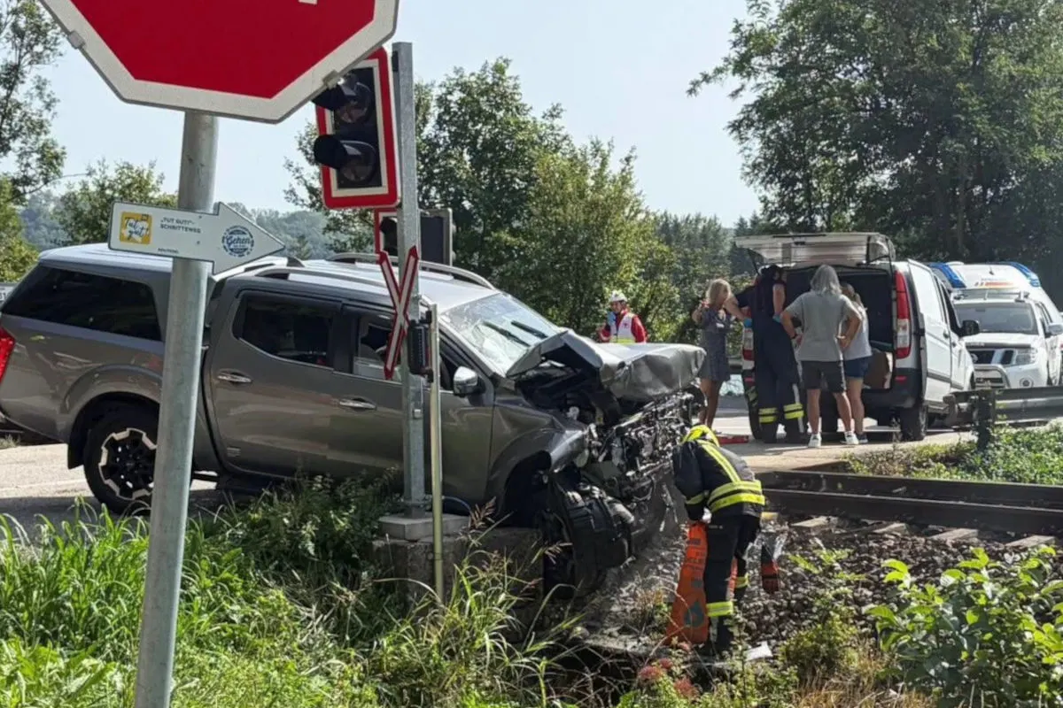 Ein silberner SUV steht stark beschädigt vor einem Verkehrsschild an einem Bahnübergang. Feuerwehrleute sind vor Ort, um den Unfall zu sichern.