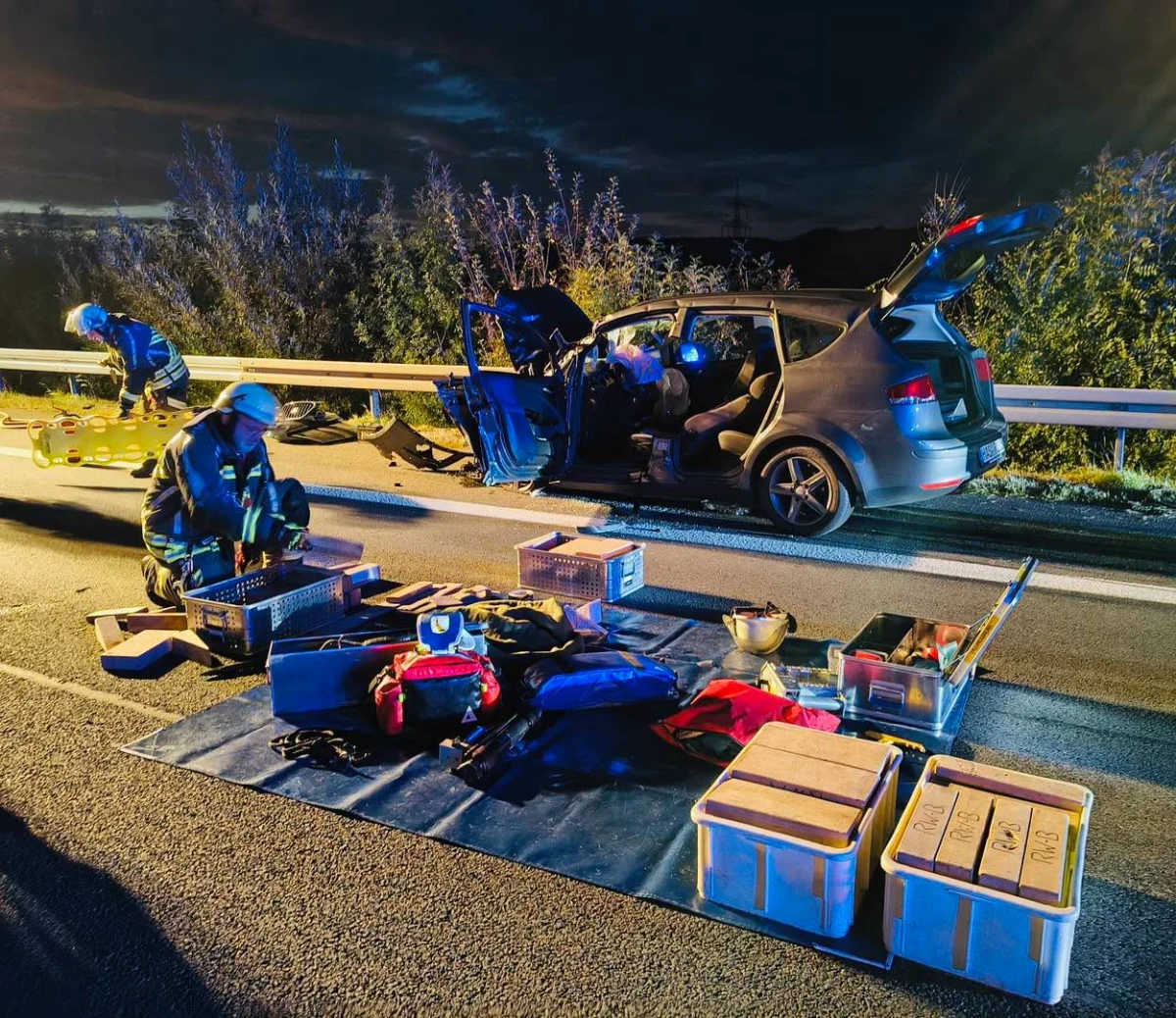 Zwei Feuerwehrleute arbeiten bei Nacht an einem verunglückten Auto, umgeben von Ausrüstung auf der Straße.