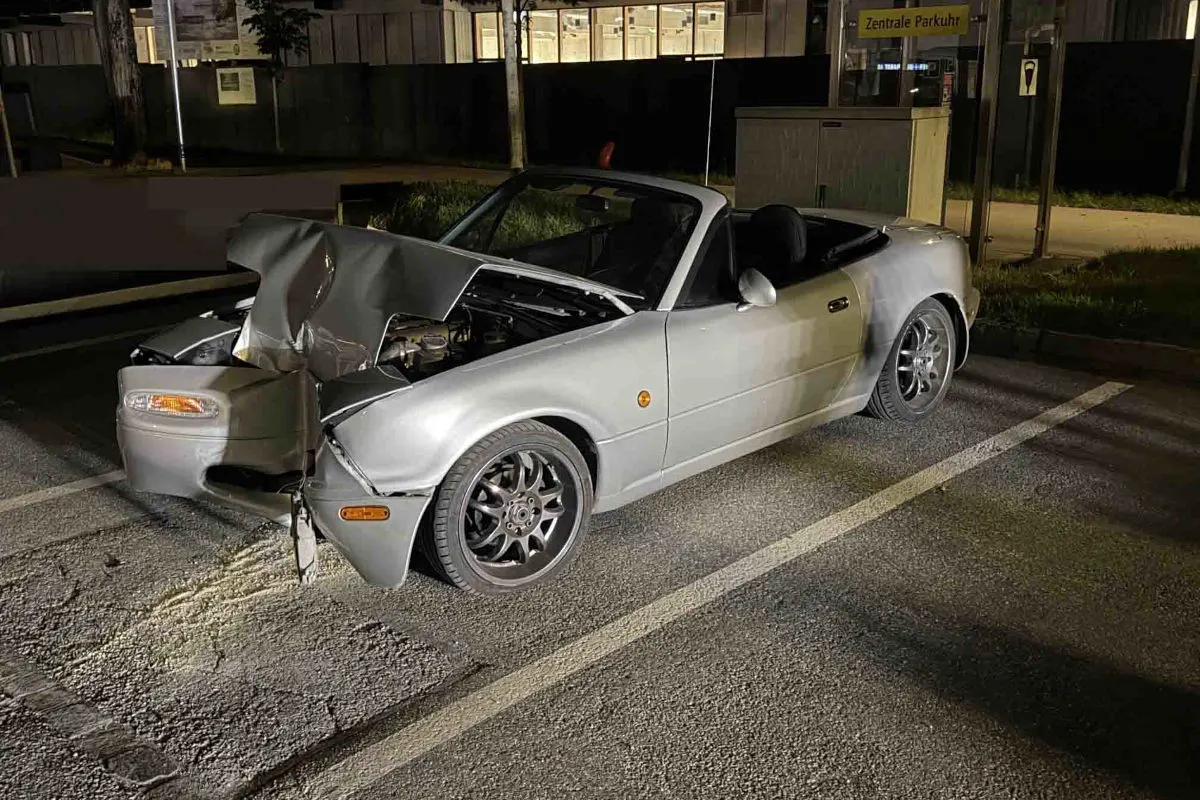 Ein silbernes Cabrio steht auf einem Parkplatz und hat einen deutlichen Frontschaden. Der Unfall ereignete sich nachts, in einer von Straßenlaternen beleuchteten Umgebung.