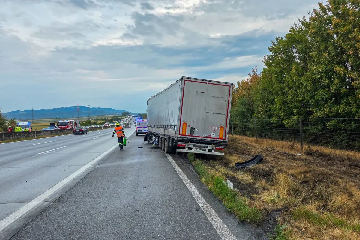 Ein Lastwagen steht halb im Straßengraben auf einer Autobahn, während ein Einsatzfahrzeug mit Blaulicht dahinter sichtbar ist. Ein Rettungsarbeiter nähert sich dem Lkw, während weitere Fahrzeuge weiter hinten zu sehen sind.