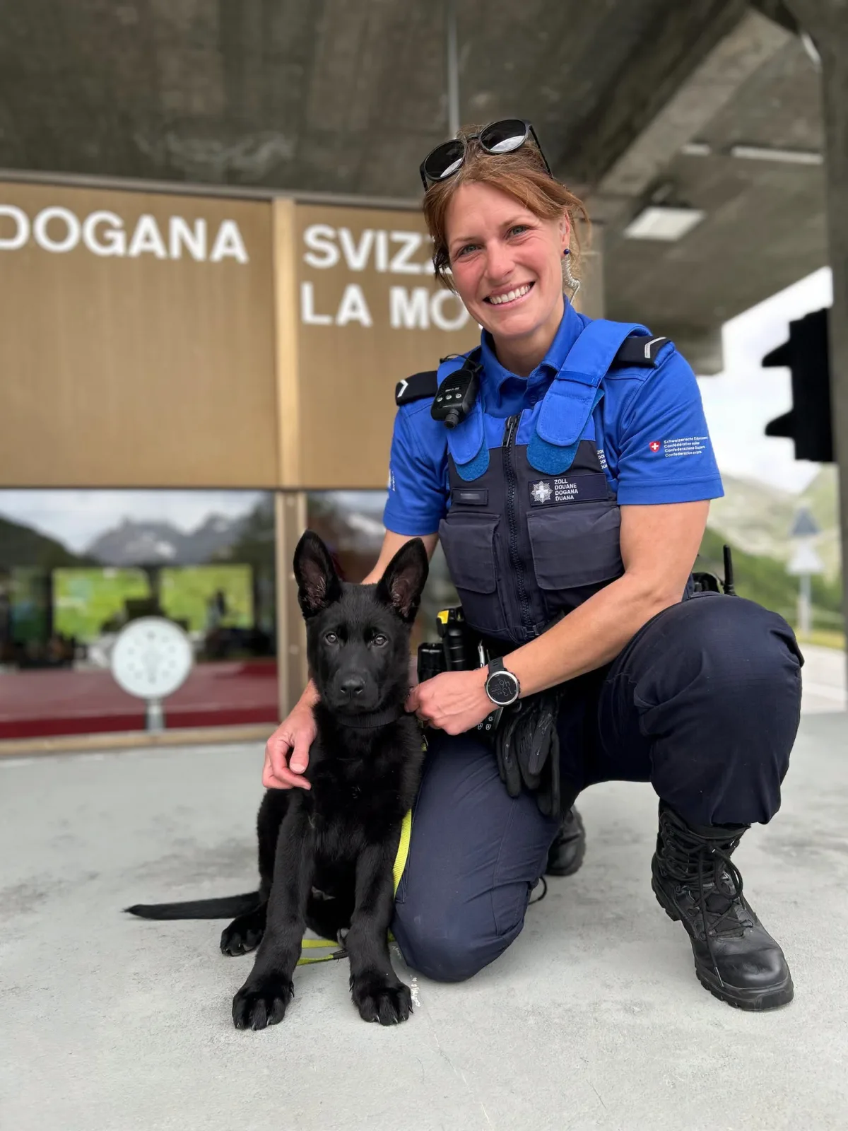 Eine Polizistin in blauer Uniform kniet vor einem Gebäude mit der Aufschrift 'Dogana Svizzera' und hält einen jungen schwarzen Schäferhund an der Leine.