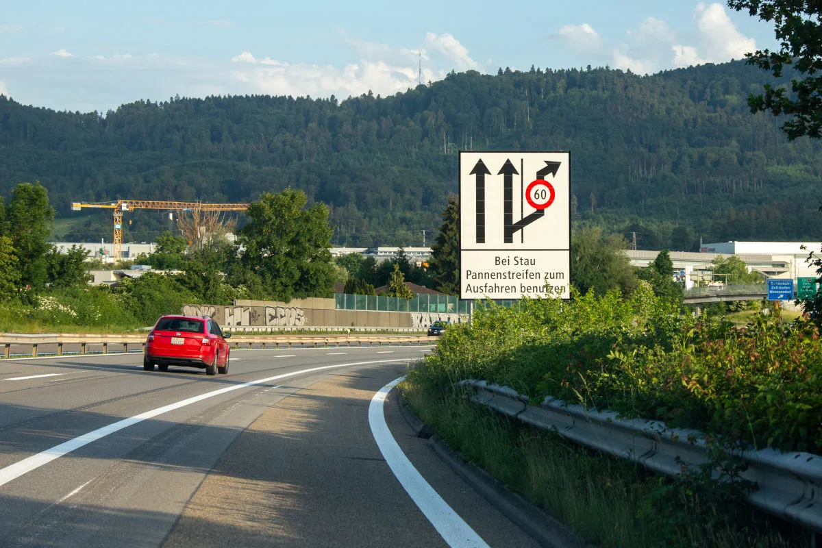 Ein rotes Auto fährt auf einer Autobahn. Rechts steht ein Verkehrsschild, das den Pannenstreifen zum Ausfahren bei Stau freigibt. Im Hintergrund sind Bäume und Hügel zu sehen.
