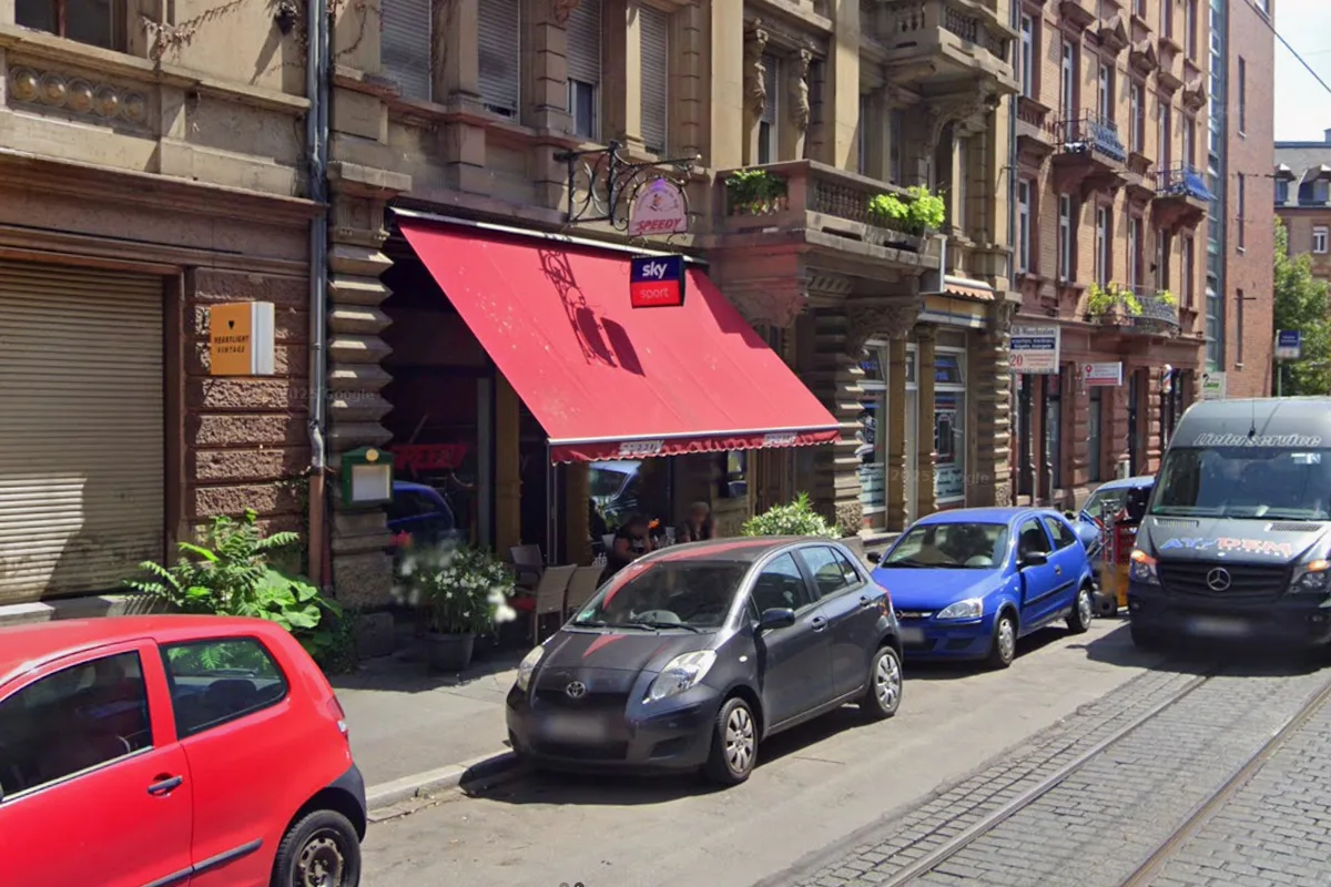 Eine Straßenszene zeigt ein Lokal mit roter Markise. Verschiedene Autos parken entlang der Straße.