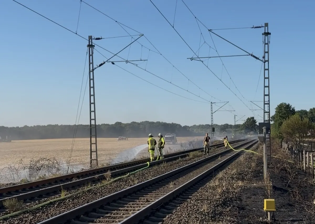 Mehrere Feuerwehrleute arbeiten auf Bahngleisen. Im Hintergrund sieht man ein Feld und Rauchwolken.