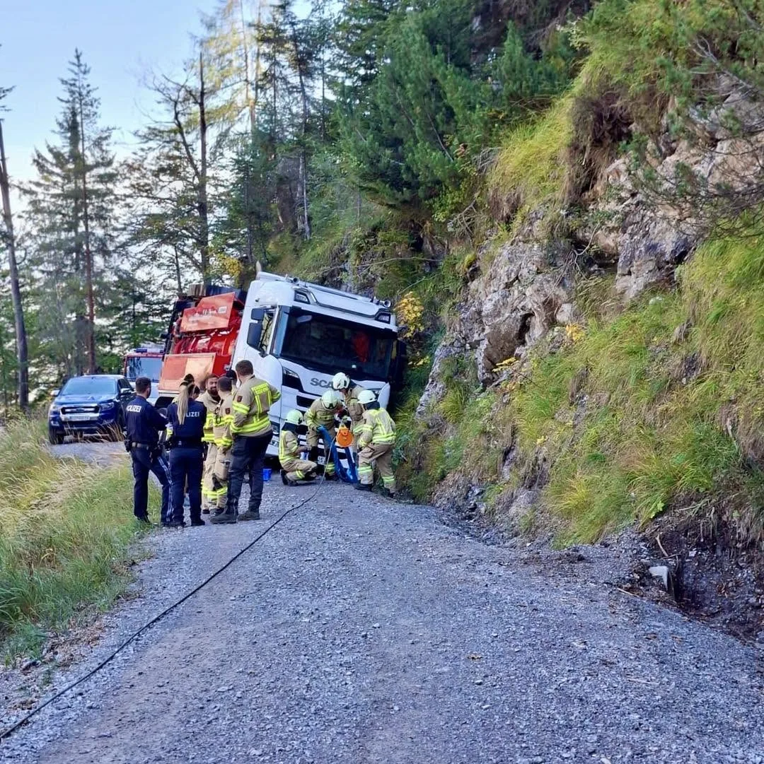 Ein Lastwagen steht schief an einem steilen, bewaldeten Hang, während Feuerwehr- und Polizeikräfte den Bereich sichern.