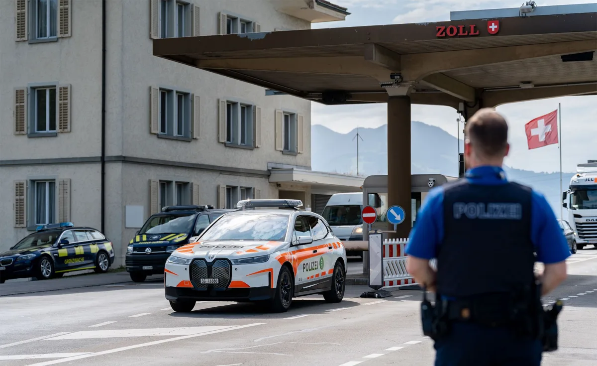 Ein Polizeifahrzeug steht vor einer Zollstation, während ein Polizist im Vordergrund sichtbar ist. Im Hintergrund weht eine Schweizer Flagge.