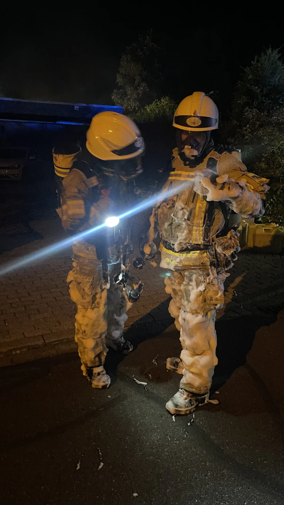 Zwei Feuerwehrleute stehen in schaumüberzogenen Schutzanzügen auf einer Straße bei Nacht. Beide tragen Helme und reflektierende Streifen sind sichtbar.