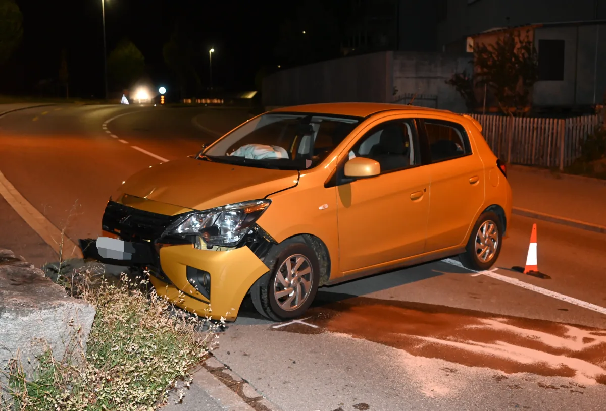 Ein orangefarbenes Auto mit sichtbaren Schäden an der Front steht auf einer Straße. Ein Verkehrshütchen und eine Kurve sind zu sehen.