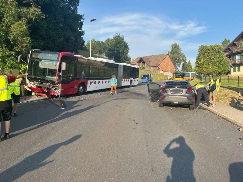 Ein beschädigter Linienbus steht nach einem Unfall auf einer Straße, neben einem schwarzen Auto. Menschen in Warnwesten sind vor Ort.