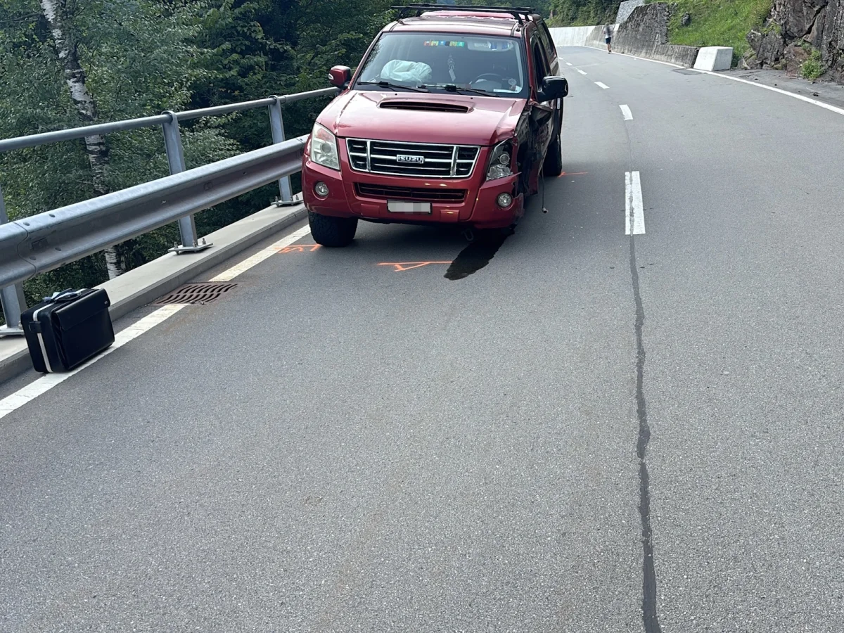 Ein roter SUV mit offensichtlichem Frontschaden steht neben einer Leitplanke auf einer kurvigen Bergstraße. Ein Koffer liegt am Straßenrand.