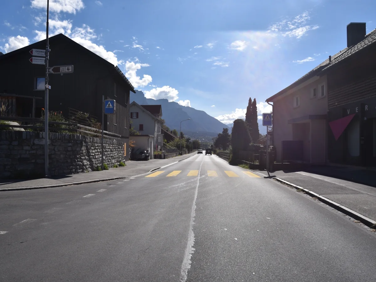 Eine leere Straße führt in Richtung der Berge. Auf der Straße sind Zebrastreifen sichtbar, am Himmel ein paar Wolken.