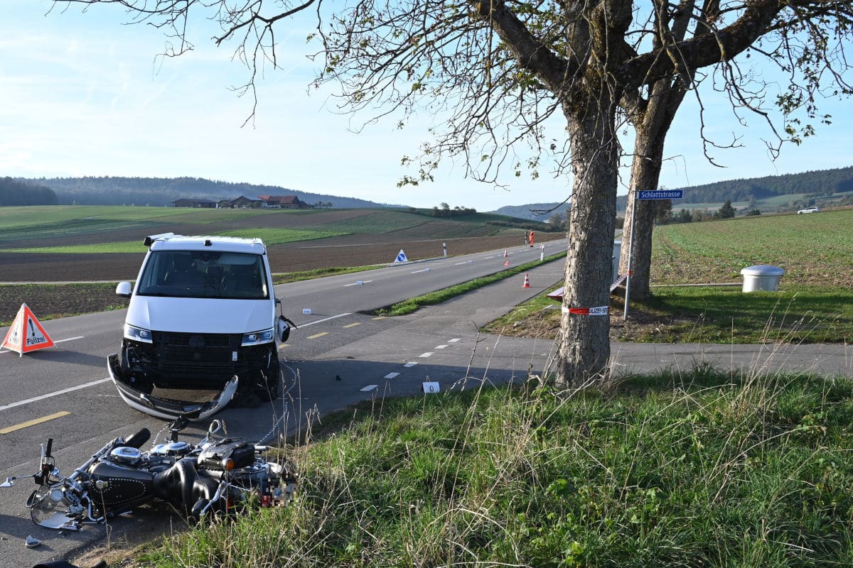 Ein weißer Van mit beschädigtem Frontbereich steht auf einer Landstraße neben einem umgestürzten Motorrad. Ein Verkehrsschild weist auf Polizeipräsenz hin.