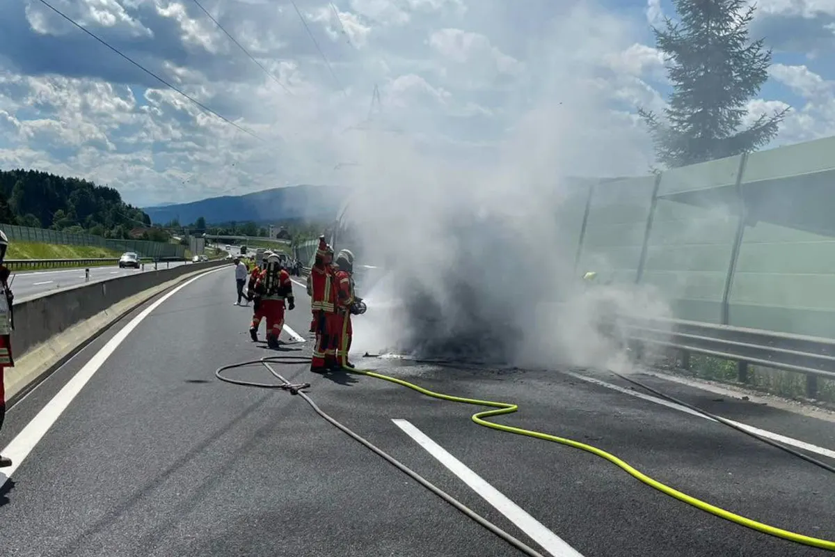 Mehrere Feuerwehrleute sind auf einer Autobahn zu sehen, während sie ein stark rauchendes Fahrzeug löschen. Der Himmel ist leicht bewölkt.