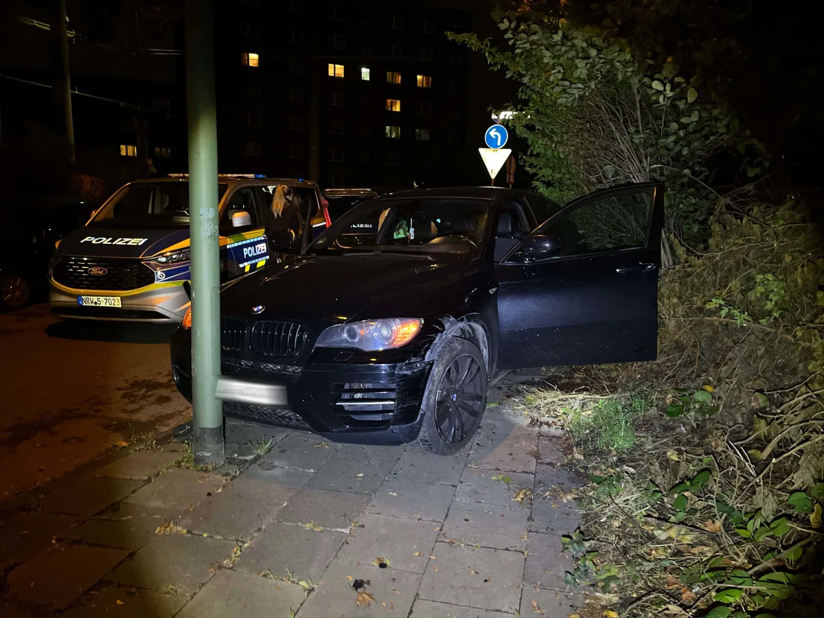 Ein schwarzer BMW mit geöffneter Fahrertür steht auf einem Gehweg. Ein Polizeifahrzeug ist in der Nähe positioniert.