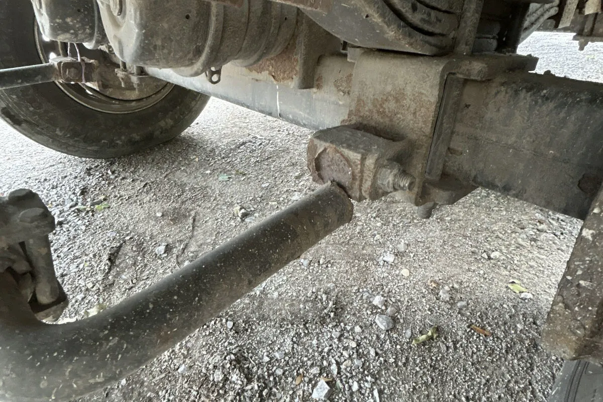 Die Nahaufnahme zeigt eine abgebrochene Metallachse unter einem Fahrzeug. Der Boden besteht aus grobem Schotter.