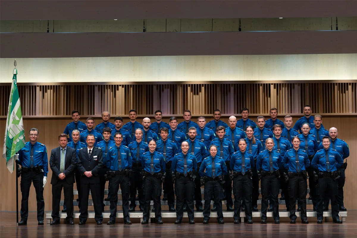 Eine Gruppe von etwa 30 Polizeibeamten steht in blauer Uniform auf Stufen für ein Foto. Sie posieren vor einem hölzernen Hintergrund, neben ihnen ist eine grüne Flagge erkennbar.