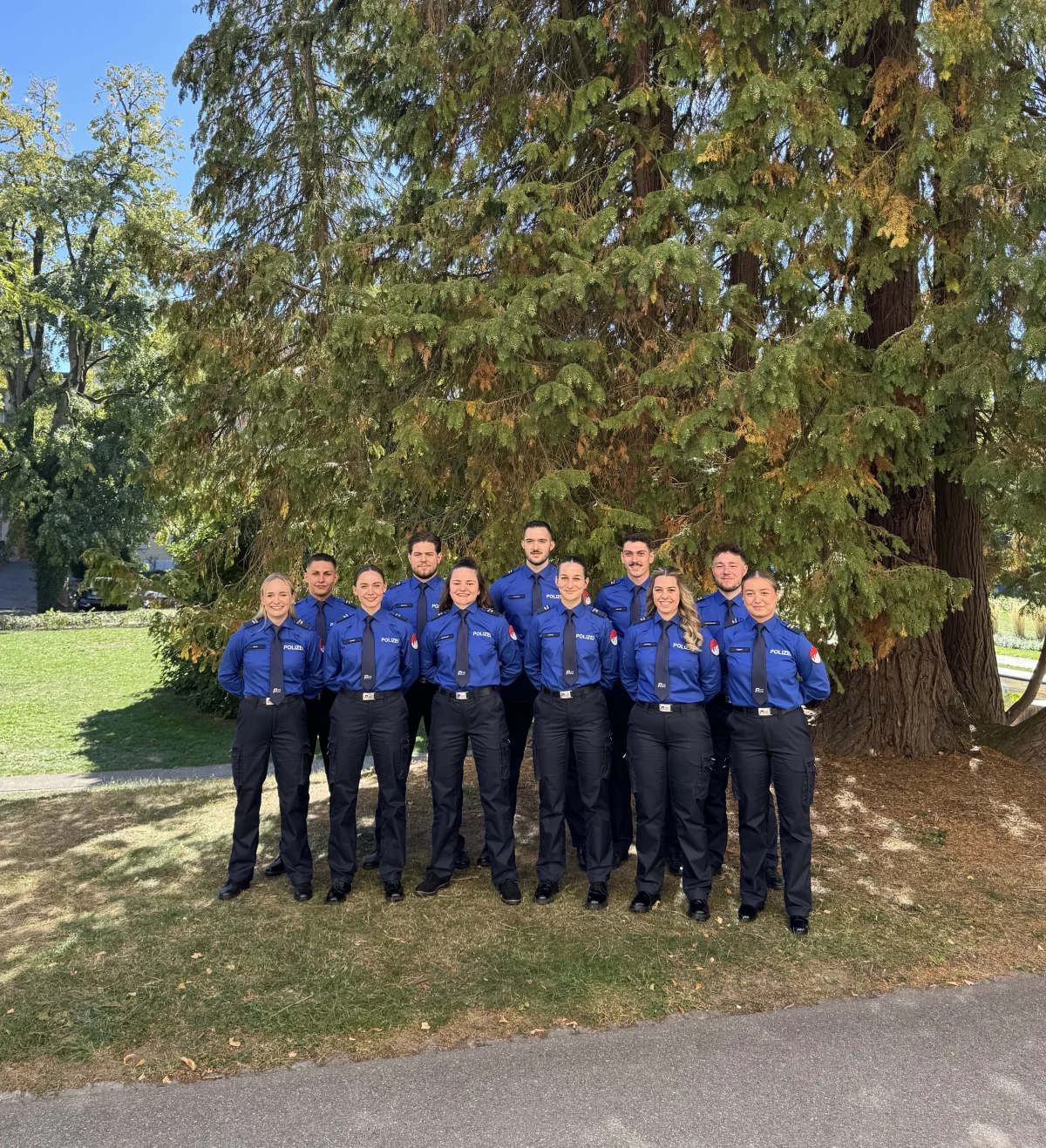 Eine Gruppe von zehn Polizisten in blauen Uniformen posiert vor einem großen Baum. Es ist ein sonniger Tag im Freien.