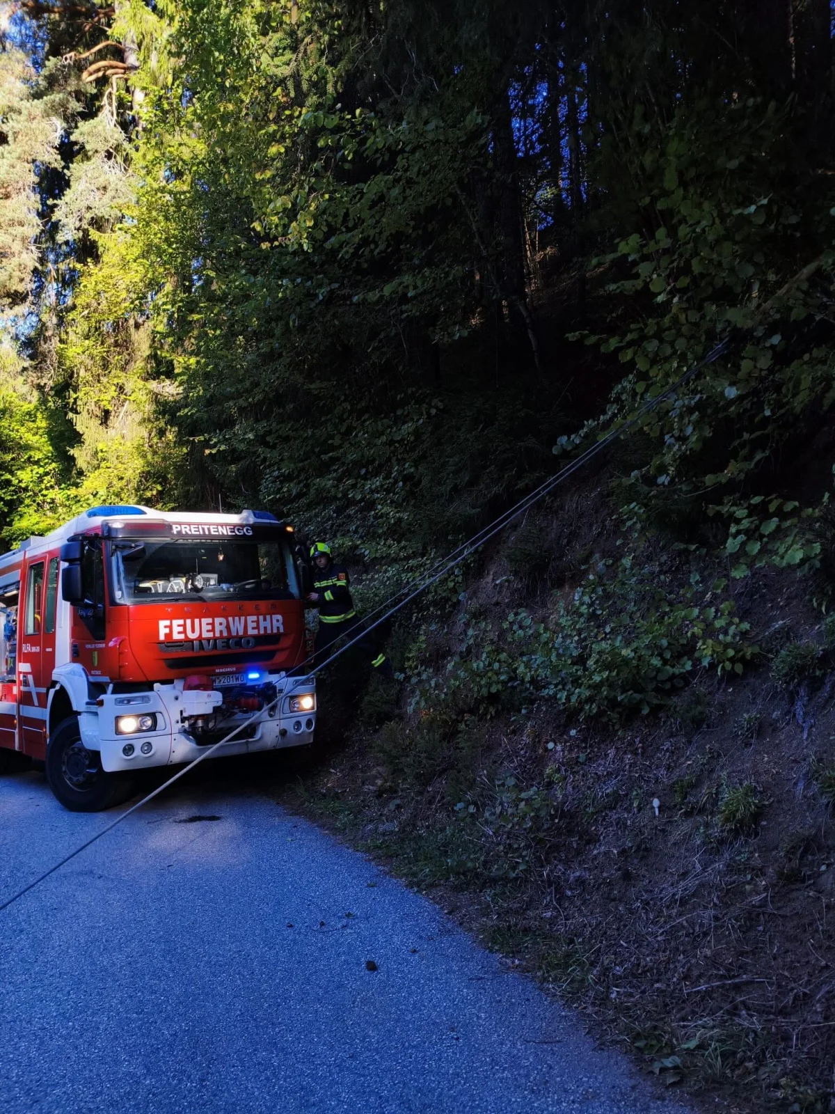 Ein rotes Feuerwehrfahrzeug der Marke Iveco steht auf einer Waldestraße und sichert mit einem Seil eine steile Böschung.