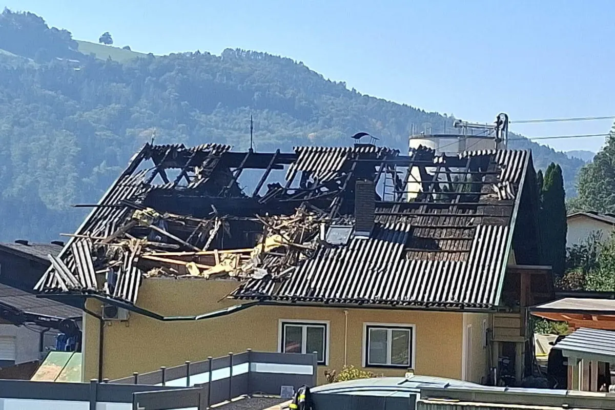 Das Bild zeigt ein Haus mit einem erheblich beschädigten Dach, das durch ein Feuer verursacht wurde. Im Hintergrund ist eine hügelige Landschaft zu sehen.