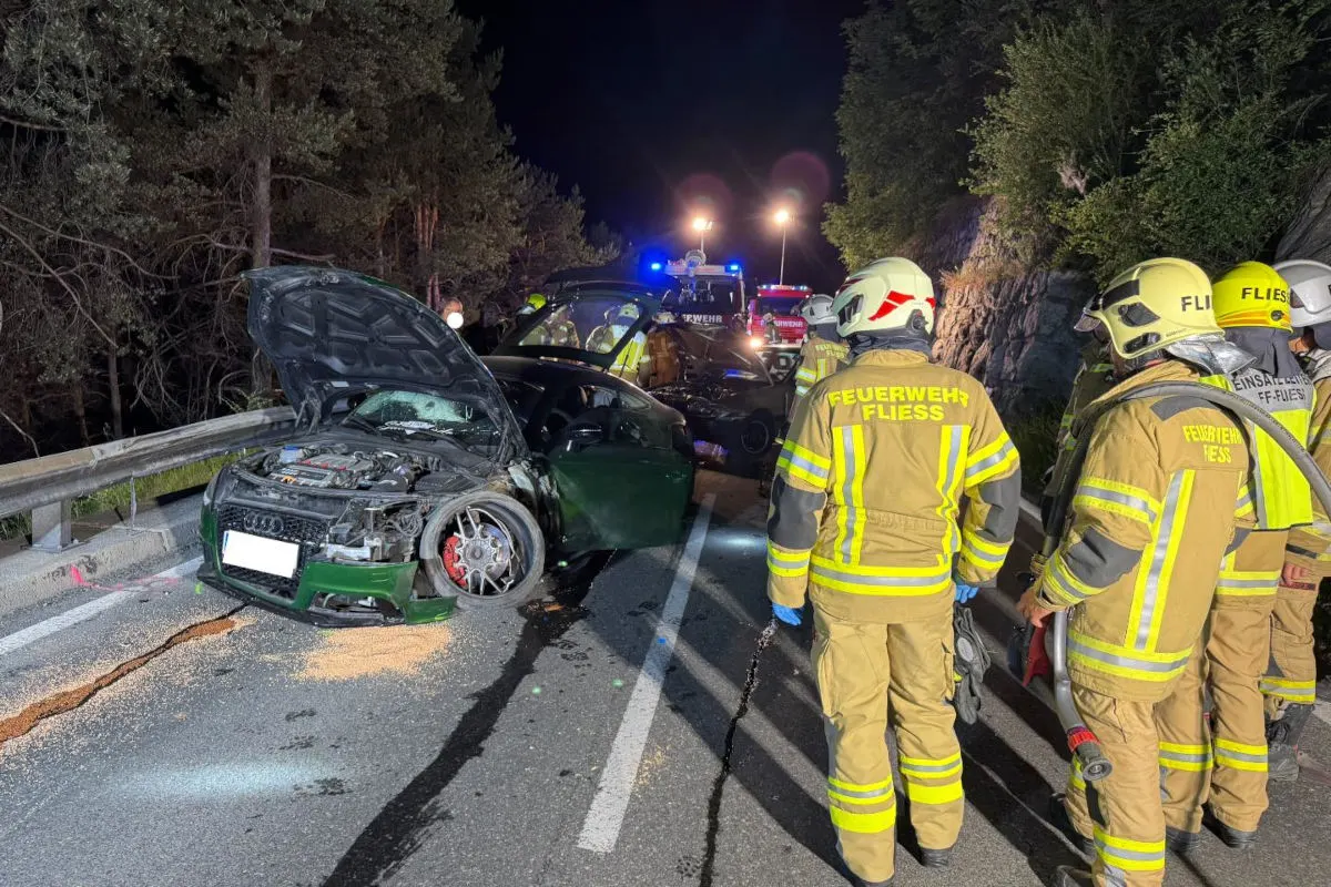 Ein beschädigter grüner Audi steht auf einer Landstraße, umringt von Feuerwehrleuten in gelben Uniformen mit der Aufschrift 'Feuerwehr Fliess'. Es ist Nacht, und weitere Einsatzfahrzeuge sind im Hintergrund sichtbar.