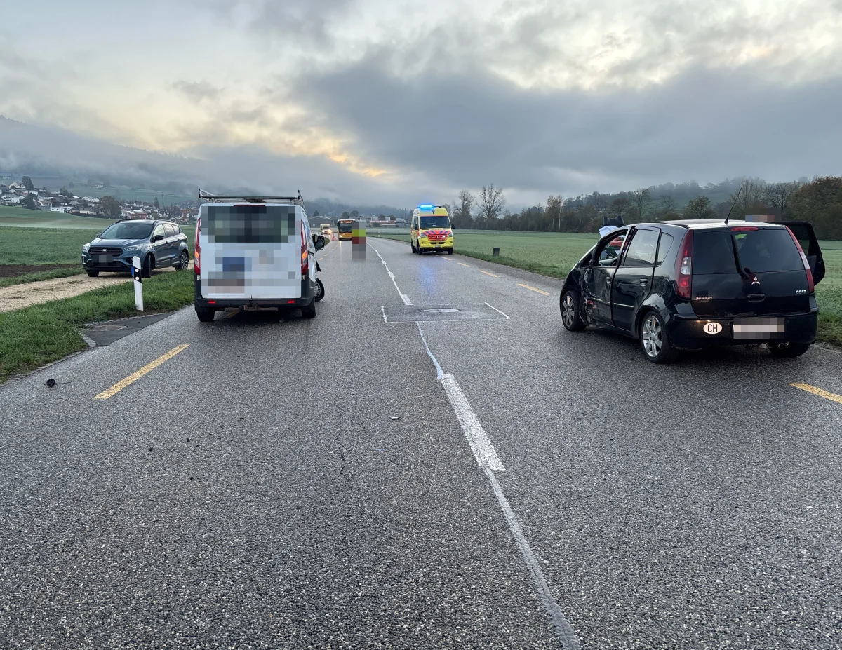 Ein beschädigtes schwarzes Auto und ein weißer Van stehen auf einer nassen, ländlichen Straße. Im Hintergrund ist ein Krankenwagen zu sehen.