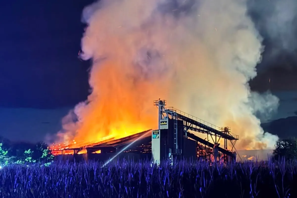Ein massives Feuer hat eine Lagerhalle erfasst, wobei Flammen und dichter Rauch sichtbar aufsteigen. Die Szene findet bei Nacht statt und wird von Notlicht beleuchtet.
