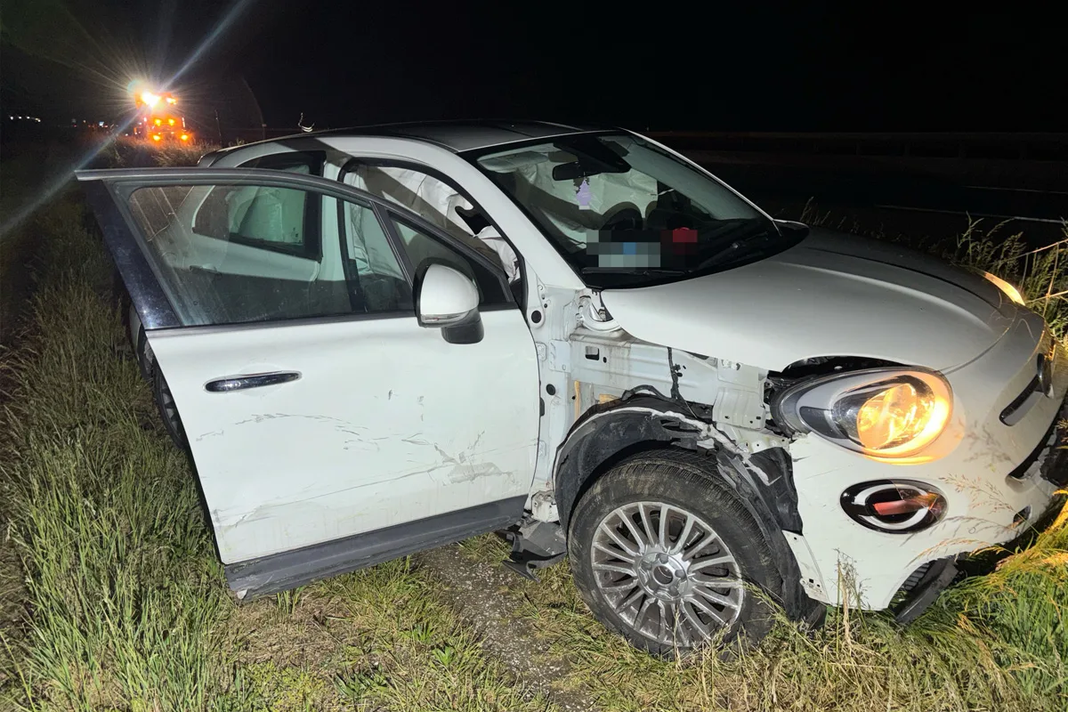 Ein weißes Auto mit deutlichen Beschädigungen steht am Straßenrand. Die Fahrertür ist offen, und die Scheinwerfer sind eingeschaltet. Im Hintergrund sind Unfallfahrzeuge zu sehen.