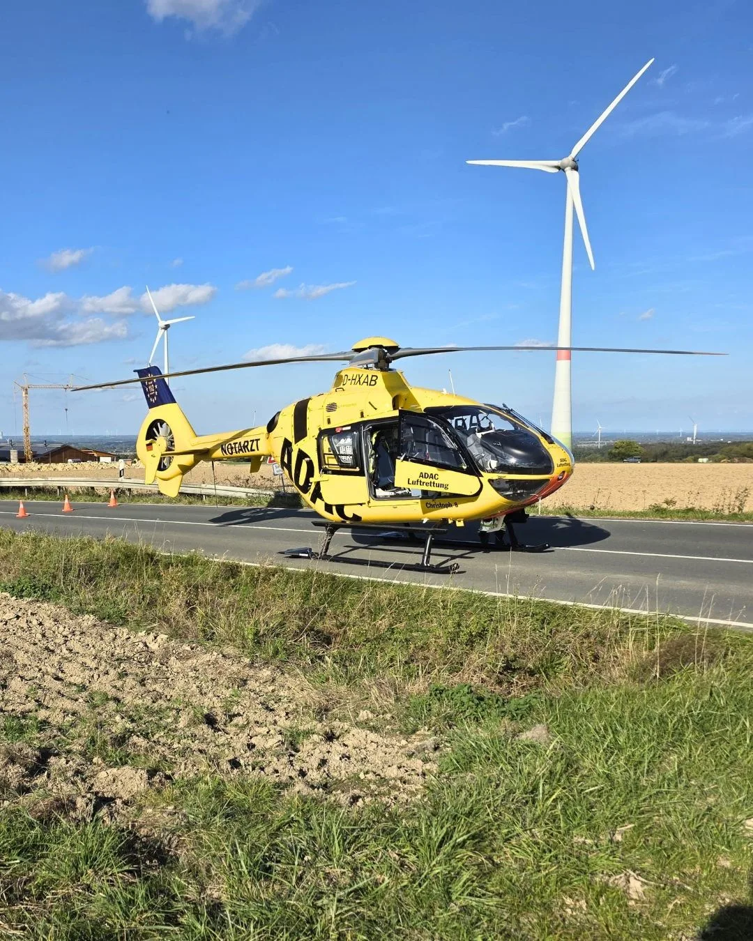 Ein gelber Rettungshubschrauber des ADAC ist auf einer Landstraße gelandet. Im Hintergrund sind zwei große Windräder und ein klarer blauer Himmel zu sehen.