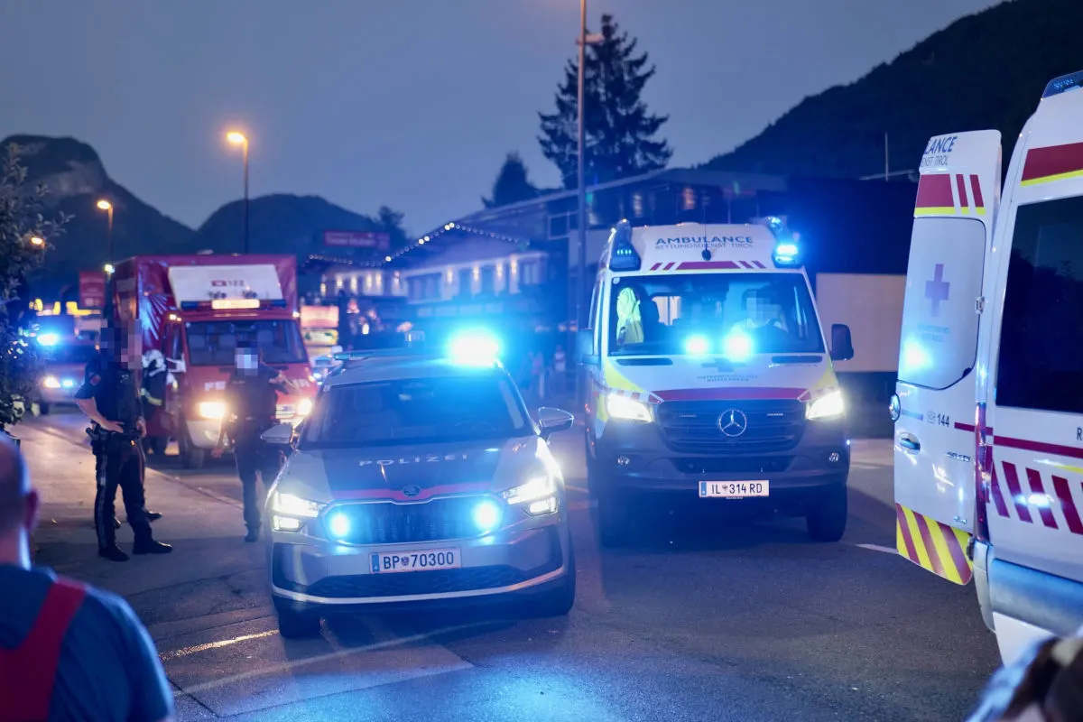 Auf einer nächtlichen Straße sind mehrere Polizei- und Rettungsfahrzeuge mit eingeschaltetem Blaulicht zu sehen. Im Hintergrund stehen Gebäude und Berge.