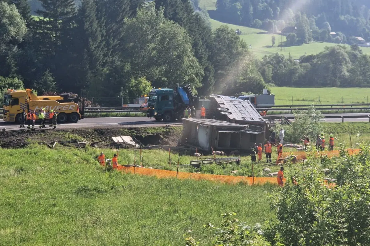 Ein Lastwagen liegt umgestürzt neben einer Autobahn. Rundherum stehen mehrere Einsatzkräfte in orangefarbenen Warnwesten. Die Umgebung ist grün, mit Bäumen und Wiesen im Hintergrund.