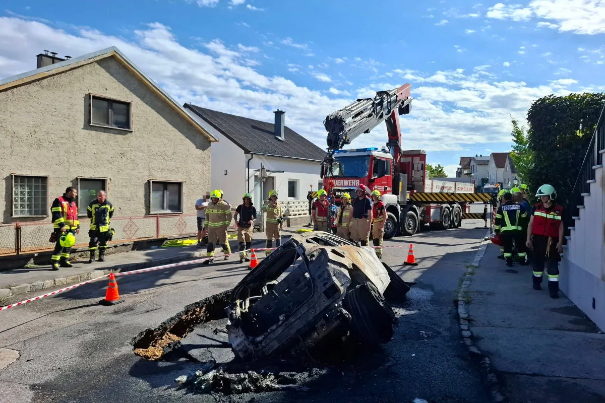 Ein zerstörtes und verbranntes Auto liegt quer auf der Straße. Mehrere Feuerwehrleute stehen in der Nähe, um die Situation zu sichern.