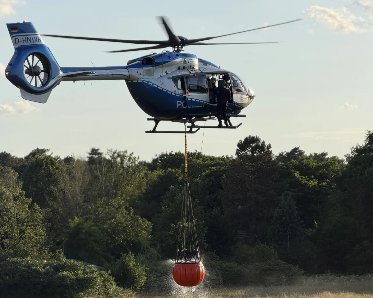 Ein blauer Polizeihubschrauber hängt einen großen, roten Wasserbehälter unter sich, während er über einem Wald schwebt. Ein Mann in Uniform ist an der Tür des Hubschraubers sichtbar.