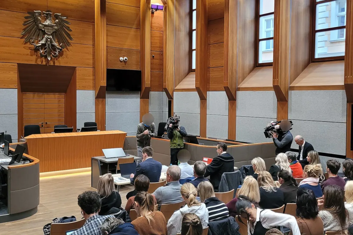 Der Gerichtssaal ist voll besetzt mit Zuschauern, während Kamerateams Aufnahmen machen. Ein großes Wappen hängt über dem Richterpult.