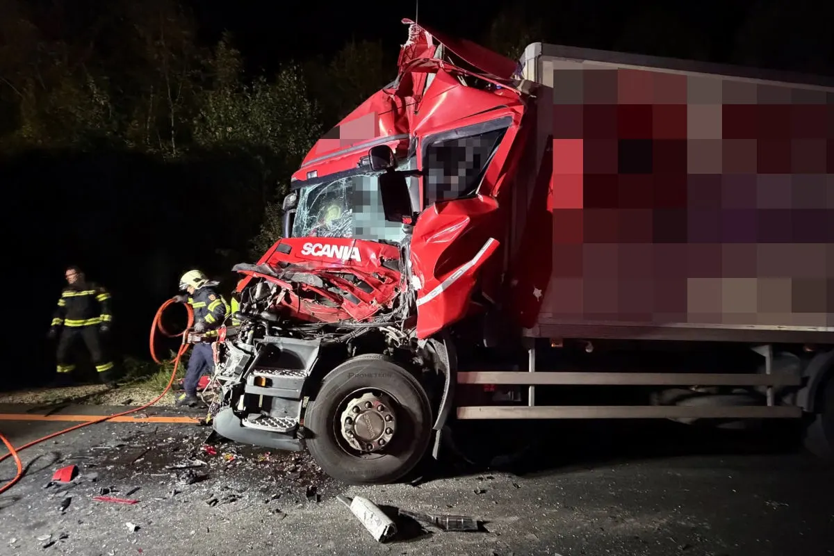 Ein stark beschädigter roter Lkw steht auf einer Straße, umgeben von Feuerwehrleuten, die Rettungsarbeiten durchführen. Die Front des Fahrzeugs ist stark deformiert, und Trümmer liegen auf dem Boden.