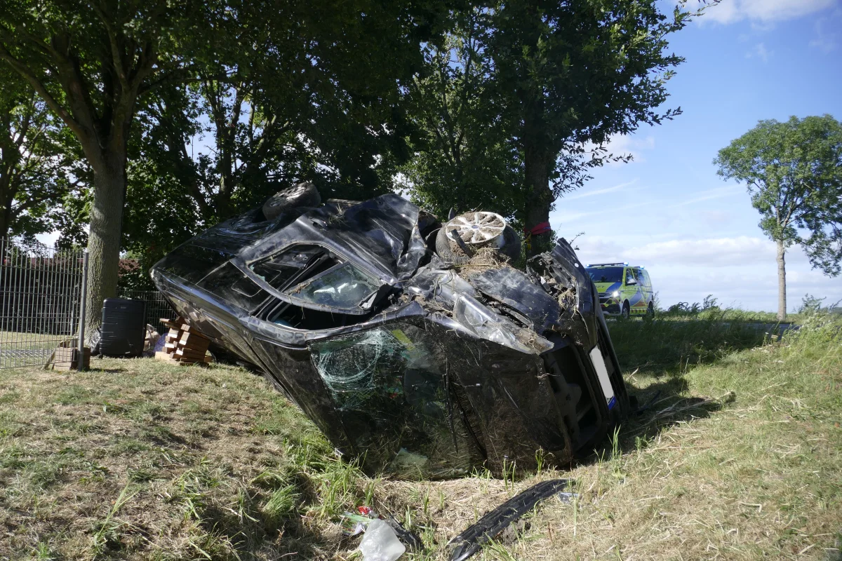Ein schwarzes Auto liegt auf der Seite neben einem Baum auf einer Wiese. Im Hintergrund ist ein Rettungswagen zu sehen.