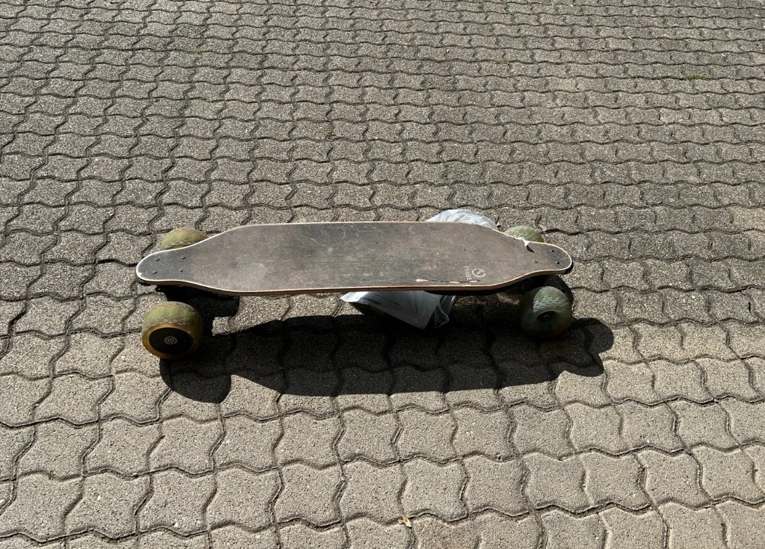 Ein Longboard mit abgenutztem Deck liegt bei Tageslicht auf einem gepflasterten Gehweg.