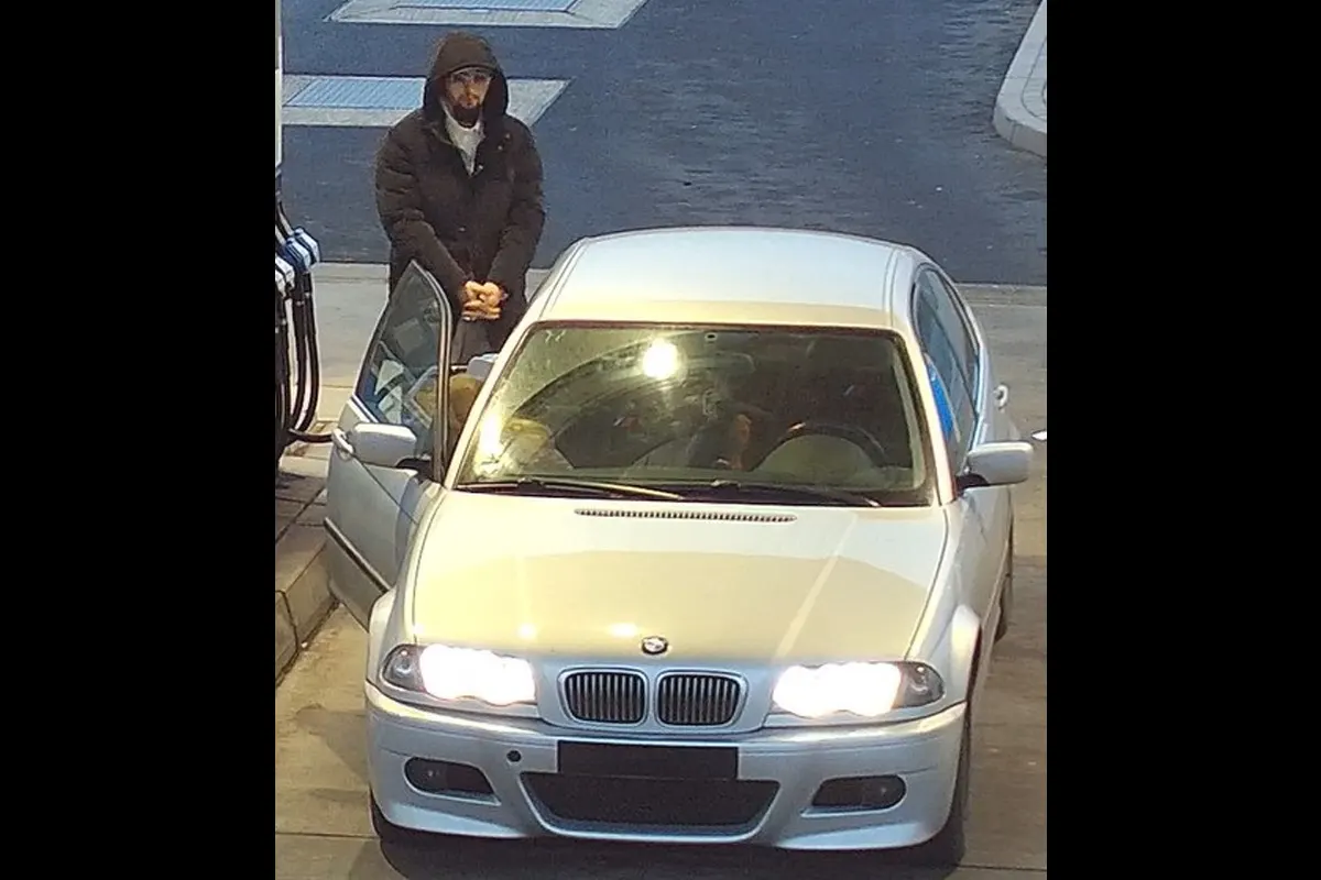 Ein Mann in dunkler Kleidung steht neben einem silbernen BMW an einer Tankstelle. Der Mann trägt eine Jacke mit Kapuze.