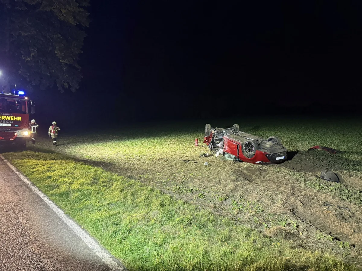 Ein rotes Auto liegt nachts überschlagen auf einem Feld. Daneben steht ein Einsatzfahrzeug mit Feuerwehrleuten.
