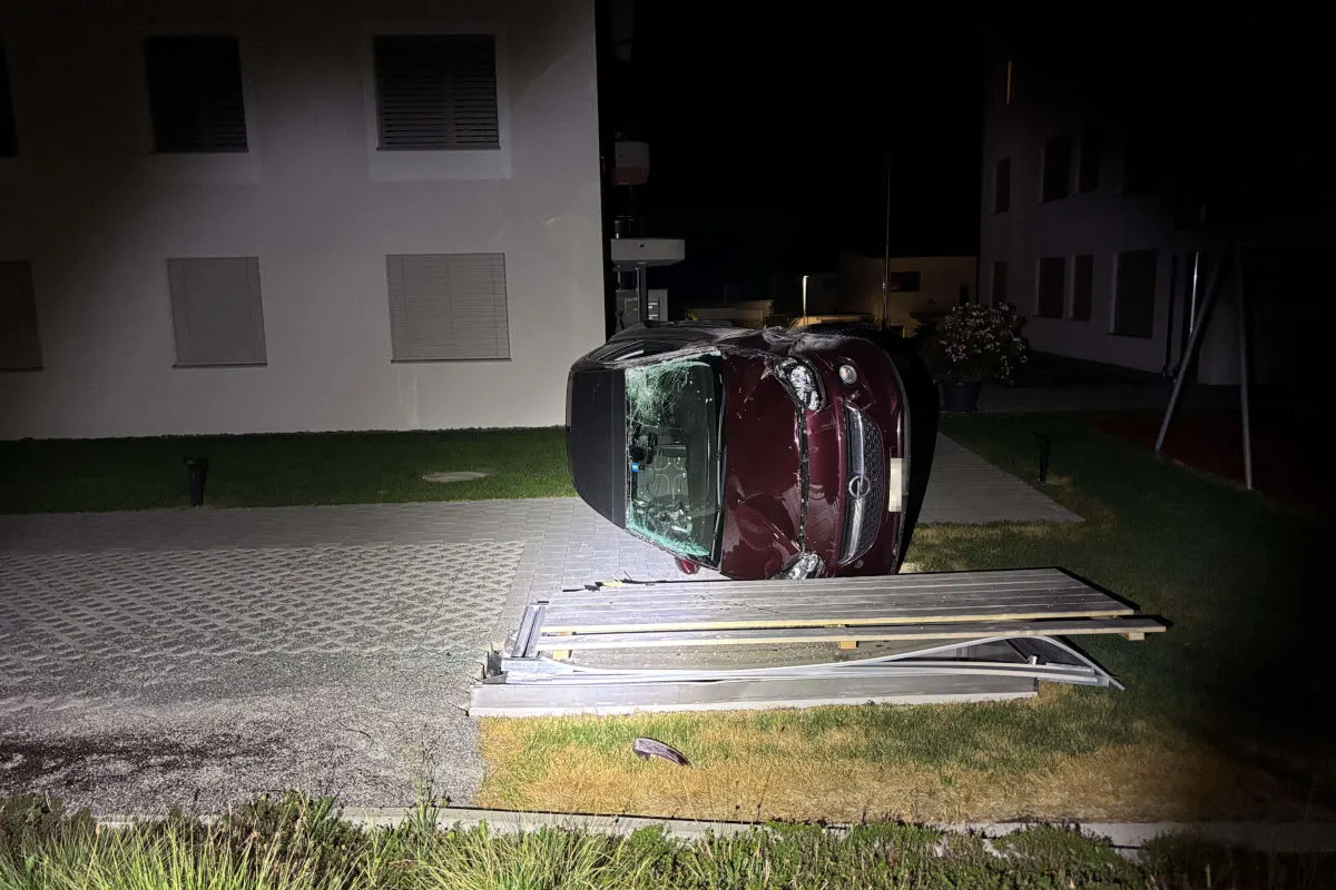 Ein Auto liegt auf der Seite vor einem Gebäude, umgeben von zerstörtem Metallzaun. Es ist Nacht.