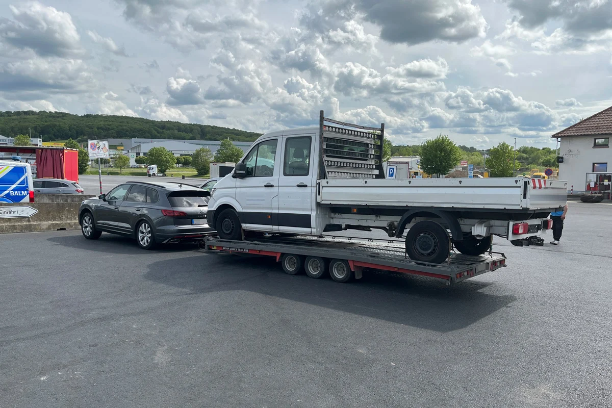Ein weißer Transporter steht auf einem Anhänger, der von einem grauen SUV gezogen wird. Im Hintergrund sind Parkplätze und ein bewölkter Himmel zu sehen.