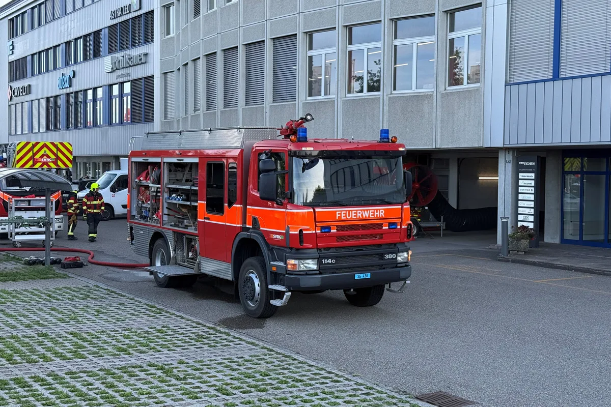 Ein rotes Feuerwehrauto mit geöffnetem Geräteraum steht vor einem mehrstöckigen Bürogebäude. Feuerwehrleute sind in der Nähe zu sehen.